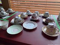 Assorted teacup and saucer sets arranged on red tabletop near window blinds.