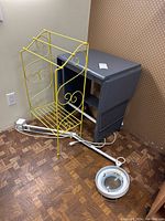 Photo showing yellow metal towel rack and gray rolling typewriter table with power strip and magnifying lamp on floor.