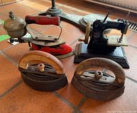 Four vintage irons and a small black miniature sewing machine arranged on a tiled floor.