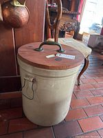 Photo showing the front and side of the vintage 10 gallon stoneware crock with wooden lid and wire handles, placed on a tiled floor.