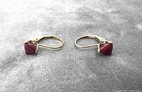 Pair of earrings laid side by side showing the red gemstones and yellow gold leverback clasps on a gray fabric background.