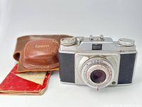 Front view of Ansco mechanical film camera with original brown leather case and instruction paper in background