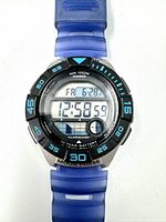 Close-up image of the Casio digital watch face showing time, day, date, and moon phase. Black and blue bezel with numeric markings around the rim and a translucent blue resin strap partially visible.