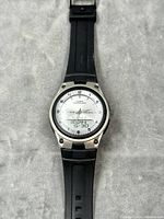 Front view of Casio AW-80 watch with white analog dial and digital display showing day, date, and time. Black resin strap visible.