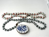 Two porcelain bead chain necklaces, one with red-orange floral beads, one with black floral beads, displayed with porcelain oval trinket box featuring blue floral design and metal trim.