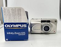 Front view of the silver Olympus Infinity Zoom 105 film camera next to its original box, showing the camera body with branding and lens cover.