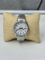 Front view of Timex Indiglo watch on burlap pillow showing white dial, black numerals, and date window.