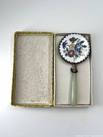 Full view of the vintage hand mirror in its original box showing the jade handle and decorative hand-painted floral back encased in a silver frame.