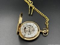 Open view of gold-toned mechanical pocket watch showing transparent dial with Roman numerals and visible internal gears. Attached chain visible.