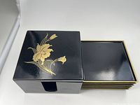 Square black lacquerware trinket box with gold floral decoration on the lid, partially open showing nested plates inside.