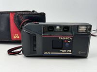 Front view of Yashica L AF camera showing lens, flash, and branding. Original case in background.