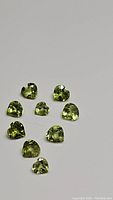 Nine heart-shaped apple green peridot gemstones arranged loosely on a grey background showing facets and clarity.