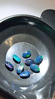 Seven oval cut calibrated black opal gemstones showing multicolored flashes on a black tray under bright light.