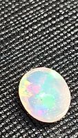 Oval cabochon Ethiopian opal gemstone lying on textured black surface showing multi-color play of color flashes under bright light.