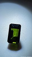 Close-up photo of a deep rich green, emerald-cut tourmaline gemstone laying on white surface, showing facets and clarity