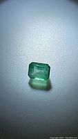 Rectangular emerald cut green gemstone (emerald) under bright light showing its color and clarity on a textured surface.