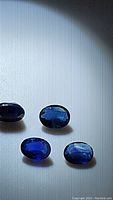 Four oval cut blue sapphires in various sizes and shades of blue placed on a white background with overhead lighting showing their faceted surfaces and deep color.