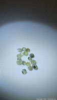 Photo of 17 small natural alexandrite gemstones with green and yellowish coloration under normal white light on white background.