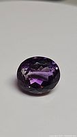 Close-up photo of a deep purple, oval cut natural amethyst gemstone on a white background. Showcases the faceted top and clarity of the stone.