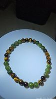 Bracelet displayed in a circular shape on white background with light focus emphasizing the green jade and tiger's eye beads' colors and patterns.
