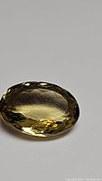 Close-up photo of a transparent golden-yellow oval citrine gemstone with faceted cut on a plain grey background.
