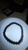 Photo of a natural black lavastone beaded stretchy bracelet placed flat on a lit white circular platform.