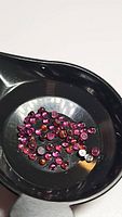 Photo of multiple small round cabochon rhodolite garnet gemstones contained within a small black scoop dish, showing their deep pink to purplish color and translucent nature.