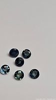 Six round cut blue-green sapphires arranged on a neutral surface showing color and clarity.