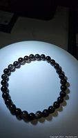 Photo of a circular stretchy bracelet made of polished round black beads with distinctive silver sheen reflections, displayed on a white lit surface.