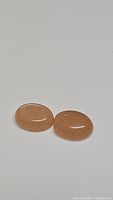 Two deep peach colored oval cabochon moonstones shown on white background, glossy smooth surfaces visible.