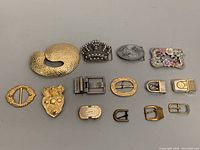 Top down view of all 14 assorted vintage metal belt buckles arranged in two rows, showcasing design variety and relative sizes.