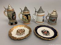 Photo showing four colorful German beer steins with pewter lids and two porcelain plates, one white and gold with crest, and one blue and gold with crest.