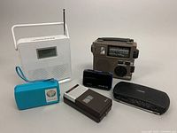 Photo showing full collection of 6 vintage electronics including portable radios, tape recorder and micro cassette player on neutral background