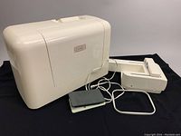 Photo of plastic cover, foot pedal, and embroidery attachment beside the machine.