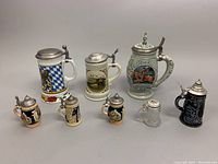 Eight vintage German beer steins and mini steins arranged side by side, showing front designs including crests, flags, and scenic paintings.