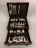 Top-down photo showing the wooden flatware chest open with knives standing vertically in the lid, rows of spoons and forks in felt-lined bottom, and serving utensils laid out.