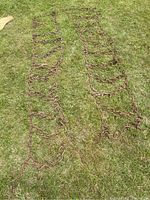 Pair of rusty metal tractor tire chains laid on grass, showing full length and zigzag pattern of links.