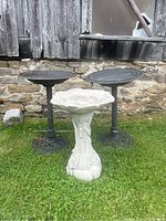 Shows three bird baths placed outdoors on grass with a stone wall background: two black metal baths and one white ornamental resin bath in front.