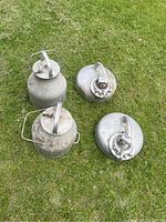 Four vintage milking pails on grass: two round Surge pails with multi-spout lids, two cylindrical De Laval pails with hinged lids, showing dirt and wear.