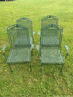 Four green metal patio chairs arranged on grass, viewed from front showing mesh seats and backs.