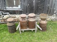 Four metal milk cans of varying sizes placed on a wooden dolly trolley on grass near a weathered wooden wall.