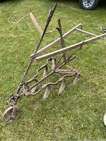 Full side view of antique walk behind plow on grass showing metal frame with three blades and wooden handles