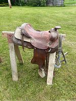 Photo showing left side of brown Western leather horse saddle on stand in outdoor grassy area, highlighting the seat, pommel, and floral tooling details.