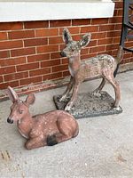 Two cement deer lawn ornaments, one standing and one lying down in front of a brick wall.