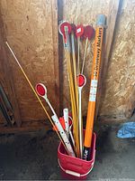Lot of multiple driveway markers with yellow poles and red circular reflectors, along with an orange storage tube, all placed inside a red bucket in a wooden shed.