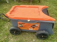Side view of the Fiskars Sit & Store gardening cart showing the orange lid, gray body, and large black wheels on grass.