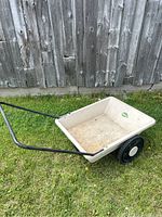 Full side view of Greenleaf wheelbarrow showing the off-white plastic bed, black metal handle, and two wheels on grass background.