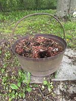 Full view of large cast iron cauldron outdoors filled with soil and leaves, showing overall shape and handle