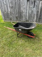 Full view of Jobmate steel wheelbarrow with black tub, orange handles and supports, resting on grass near wooden wall.