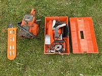 Oleo-Mac chainsaw body, detached blade guard, and Black & Decker grinder with accessories inside orange case, all placed on grass.